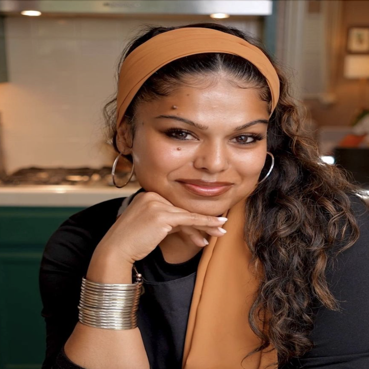 A Tasty Creator, Ronica, with long, wavy hair and a headband is smiling. She is wearing hoop earrings and stacked bracelets, resting her chin on her hand in a kitchen